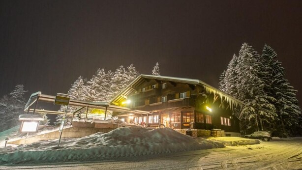 Un edificio cubierto de nieve con techo inclinado, rodeado de árboles y un montículo de nieve.