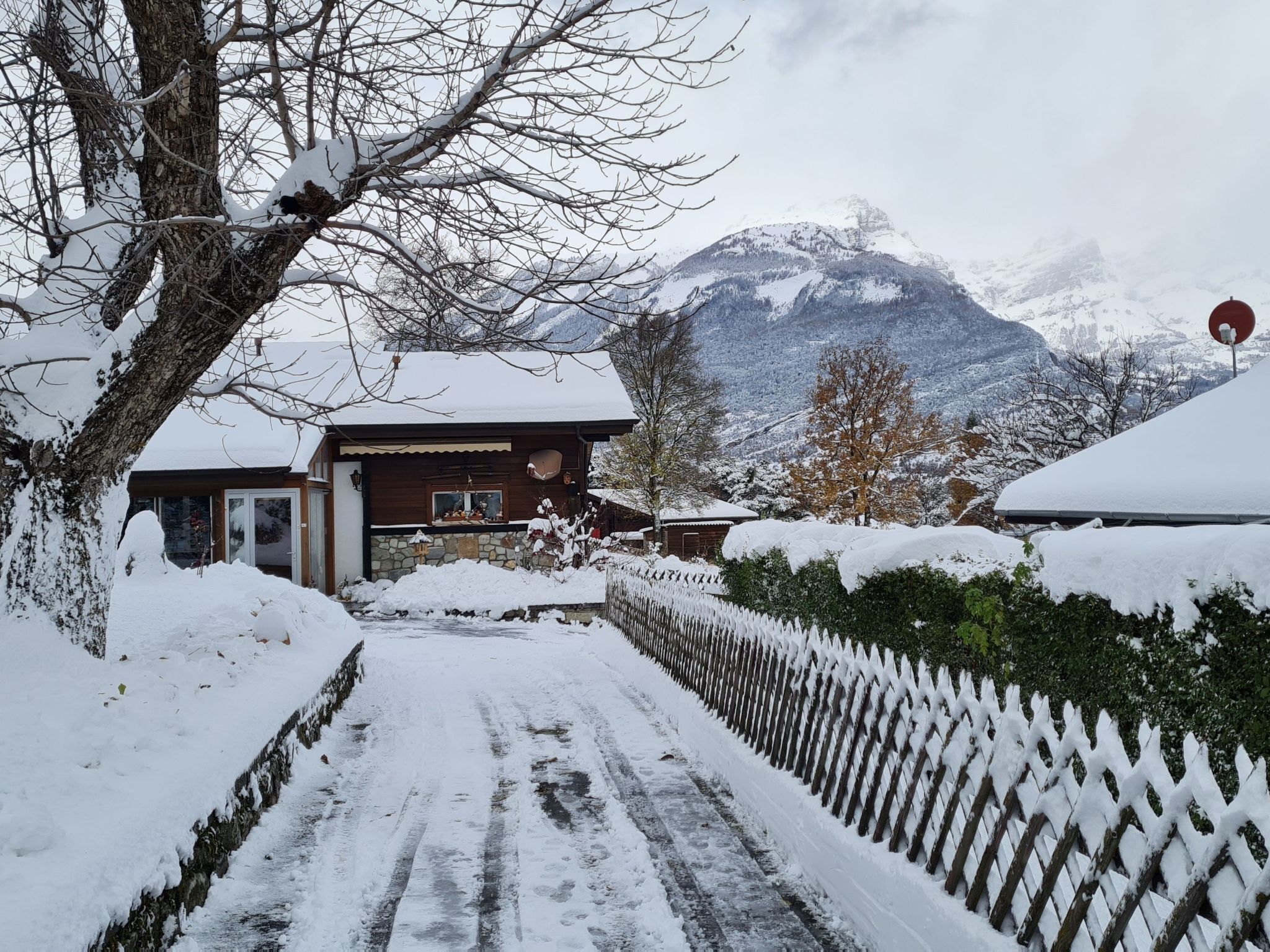 A snow-covered pathway leading to a wooden building with a stone foundation, surrounded by a snow-covered fence and trees.