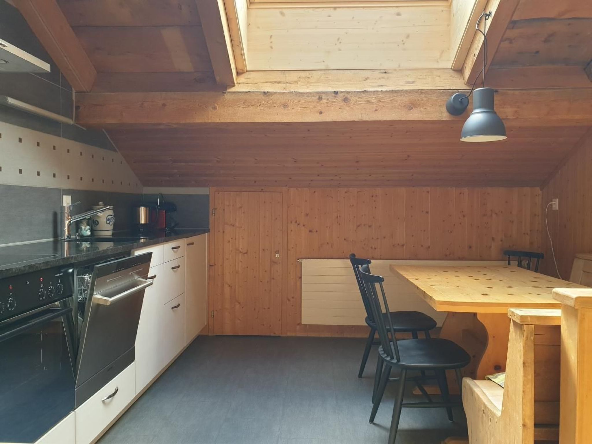 Une cuisine compacte aux murs en bois, dotée d’un plan de travail noir, d’une table à manger en bois avec deux chaises, et d’une lucarne au plafond.