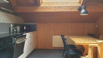 Une cuisine compacte aux murs en bois, dotée d’un plan de travail noir, d’une table à manger en bois avec deux chaises, et d’une lucarne au plafond.