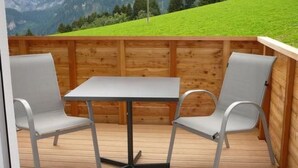 Een houten terras met een tafel en twee stoelen, met uitzicht op een groene heuvel en bergen.
