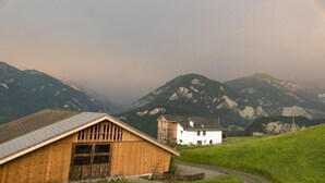 Een houten schuur en een wit huis met een berg op de achtergrond en een regenboog.