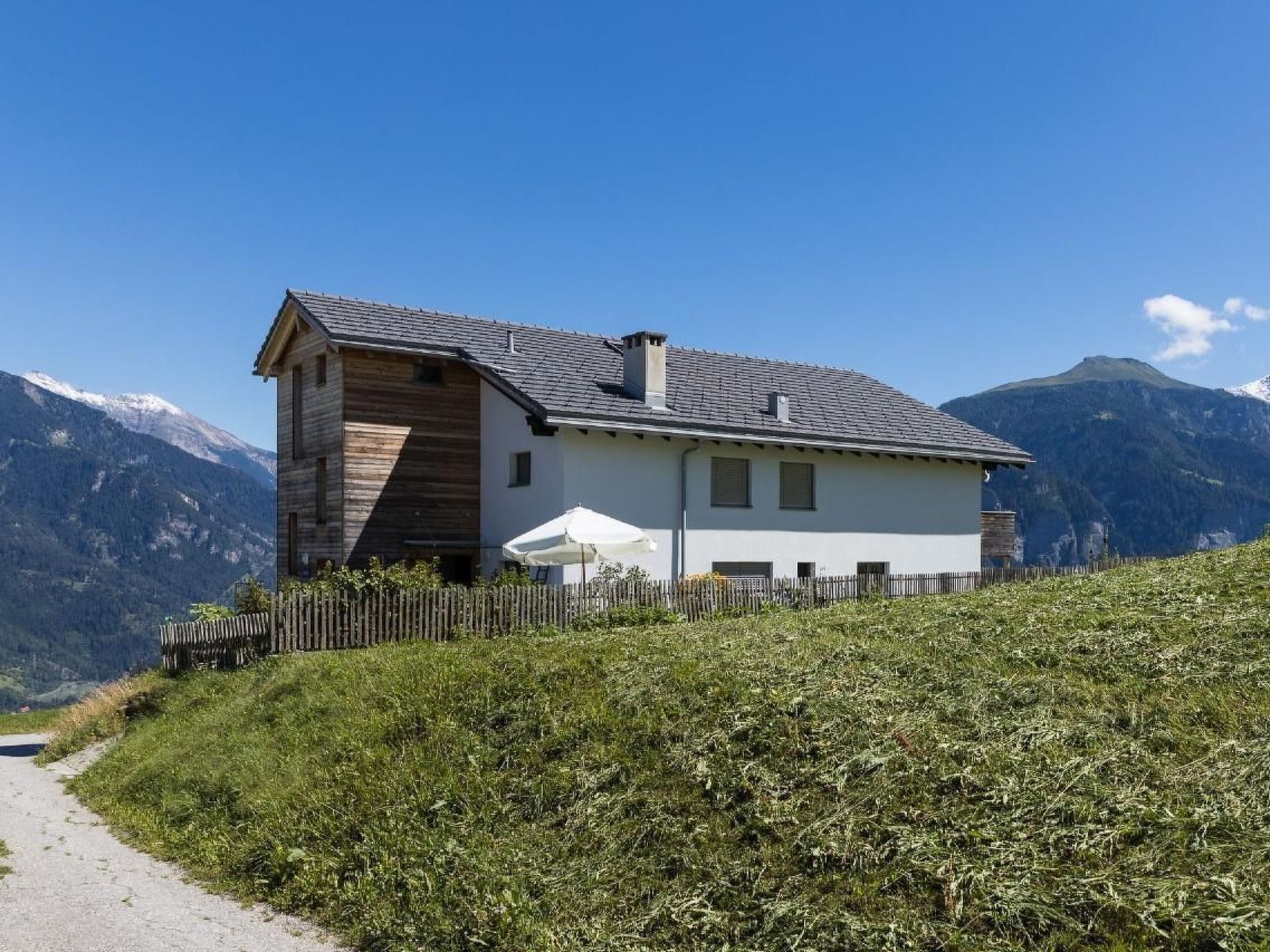 A two-story house with a sloped roof, wooden exterior, and a white facade. It is situated on a hillside with a clear view of a mountain range in the background.