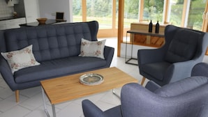 A modern living room with a grey sofa, two matching armchairs, a wooden coffee table, and a view of a kitchen and outdoor area through large windows. - "Ferienwohnung ""Spel Rein"" Cumpadials" by Interhome (Cumpadials)
