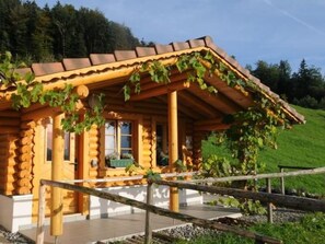 Eine rustikale Holzhütte mit geneigtem Dach, Blockbauweise und einer Laube mit Weinranken.