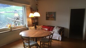 Un comedor acogedor con una mesa redonda de madera y sillas, un sofá con cojines rojos y vistas a un lago a través de un ventanal grande.