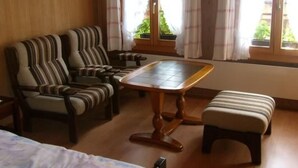 Een kamer met een bed, een gestreept bankje, een houten tafel en een raam met gordijnen.