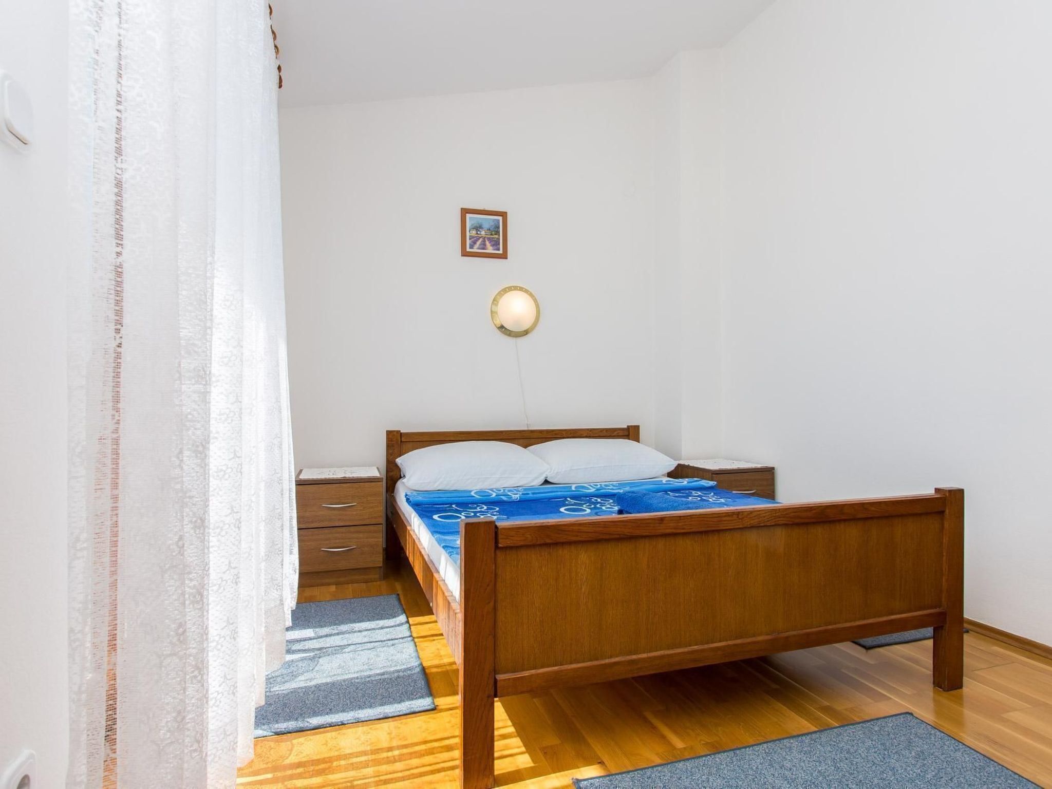 A bedroom with a wooden bed, a nightstand, a drawer, a wall-mounted light, and a framed picture.