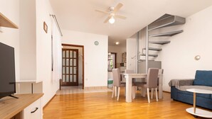 A modern living room with a wooden floor, a blue sofa, a dining area with a table and chairs, and a TV on a wooden stand. - Ceković 2 by Interhome (Krk/Šilo)