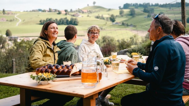A group of people sitting at a wooden picnic table outdoors with a scenic view of rolling hills in the background. - Lamahof Tschäppel by Interhome (Huttwil)