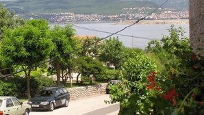Una strada residenziale con auto parcheggiate, vegetazione rigogliosa e vista su un corpo d'acqua e montagne in lontananza.