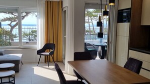 A modern living room with a wooden dining table, black chairs, and a sofa. A large window offers a view of the sea and trees. - Apartment Punta Silo by Interhome (Krk/Šilo)