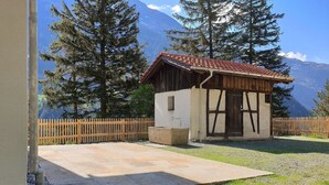 Une petite cabane en bois, surmontée d’un toit de tuiles, entourée d’une clôture en bois, et se dressant sur un fond de grands arbres et de montagnes.