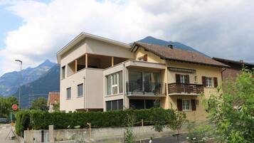 Ein zweigeschossiges Wohngebäude mit einem Balkon im oberen Stockwerk, umgeben von viel Grün und Bergen im Hintergrund.