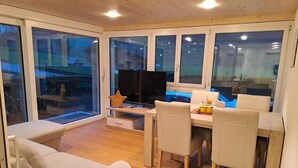 A modern dining area with a wooden table, white chairs, and a television on a stand. - Ferienwohnung by Interhome (Attinghausen)