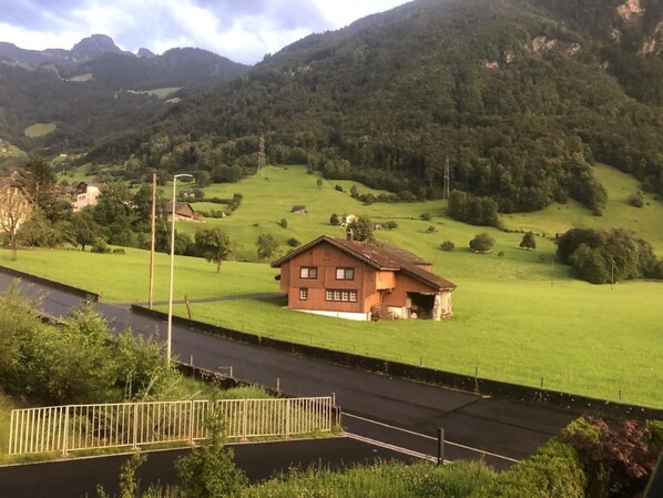 Et landhus med et skråt tag, omgivet af grønne marker og bjerge i baggrunden.