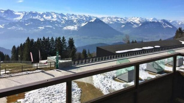 Uitzicht vanaf een balkon met glazen balustrade op besneeuwde bergen en een heldere lucht.