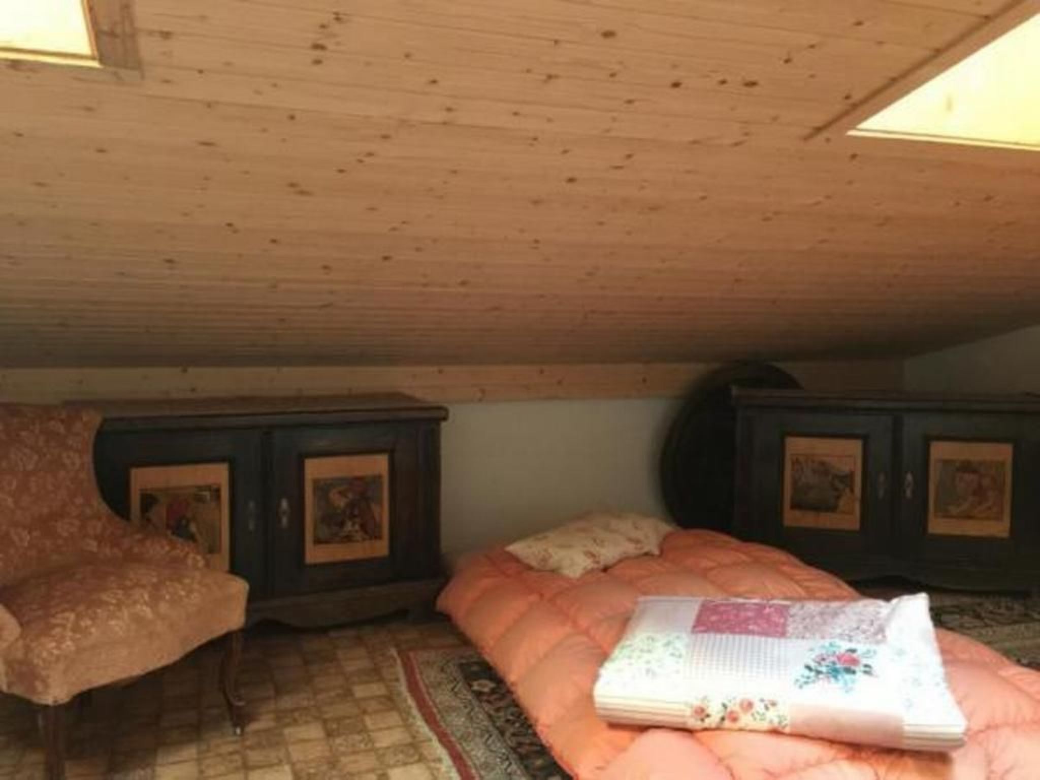 Ein gemütliches Schlafzimmer unter dem Dach mit Holzbalkendecke, einem weichen rosa Sofa, einem gesteppten Bett und zwei dunklen Holzschränken mit gerahmten Bildern.