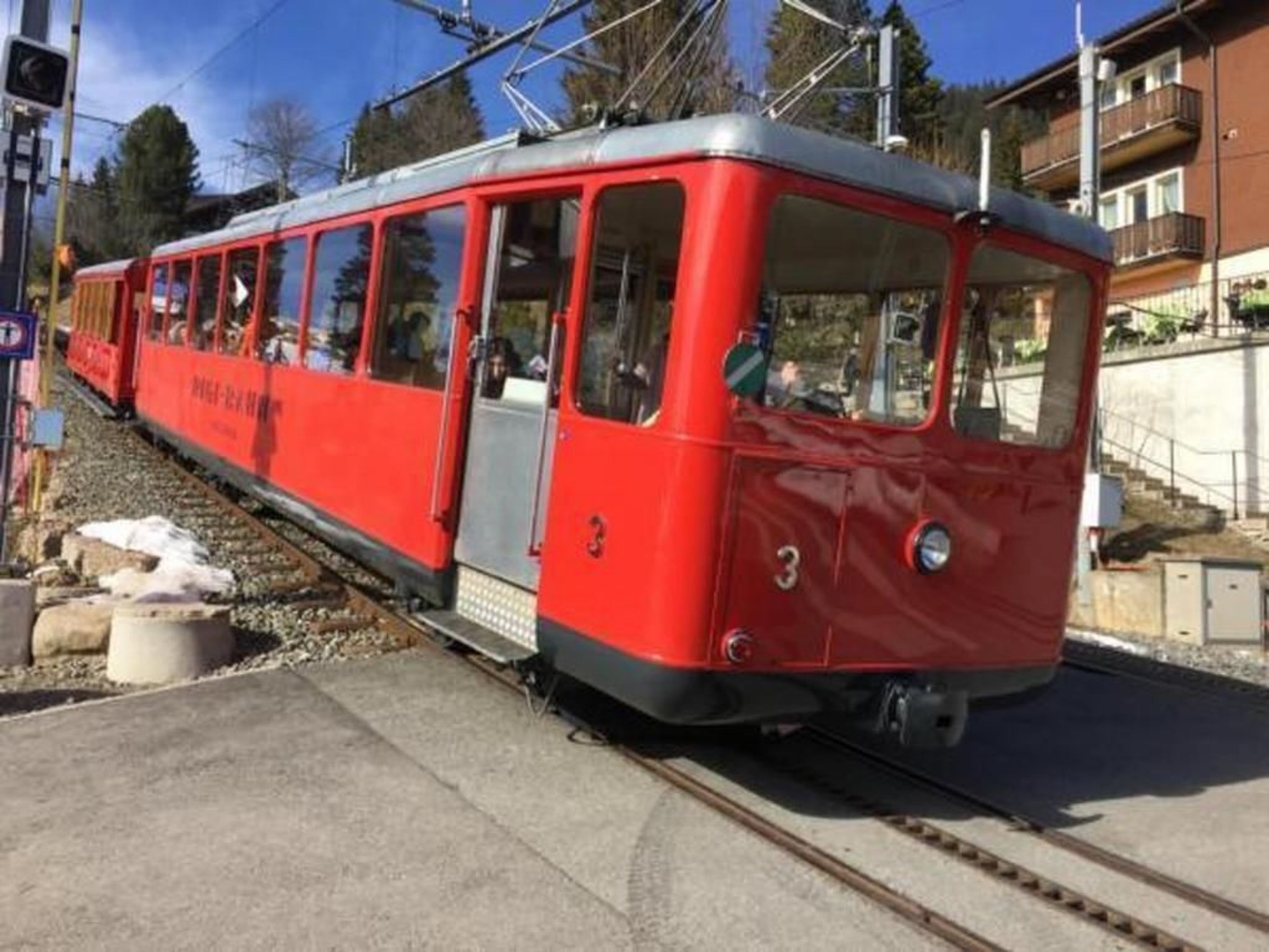 Un tramway rouge circule sur des rails, avec le numéro « 3 » peint sur le côté, et passe devant un immeuble doté de balcons.