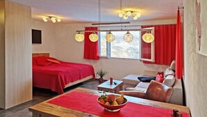 A modern hotel room with a bed, a sofa, a dining table with a red table runner, and a window with red curtains. - Studio Flumserberg by Interhome (Flumserberg Tannenheim)