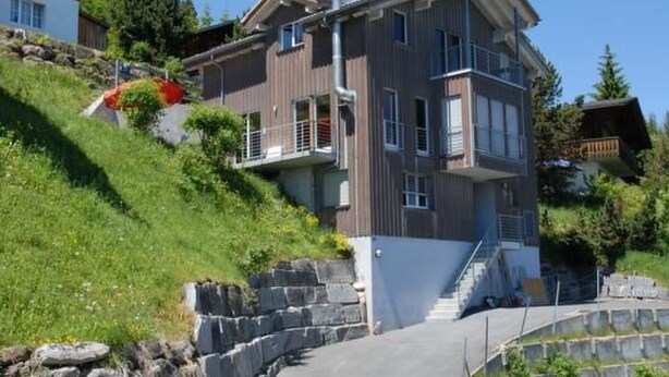 A multi-story building with a stone staircase leading to a balcony, surrounded by greenery and other buildings. - Studio Flumserberg by Interhome (Flumserberg Tannenheim)