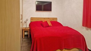 A bedroom with a wooden bed, red bedding, a small bedside table with a lamp, and a TV mounted on the wall. - Studio Flumserberg by Interhome (Flumserberg Tannenheim)
