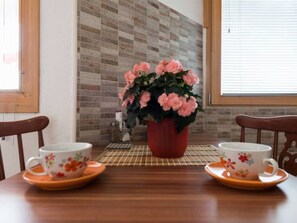 Ein Esstisch mit zwei floralen Tassen und Untertassen, einem roten Topf mit rosafarbenen Blumen und einer karierten Tischmatte.