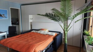 Ein modernes Schlafzimmer mit Bett, Schreibtisch und Stuhl. In der Ecke steht eine große Pflanze.