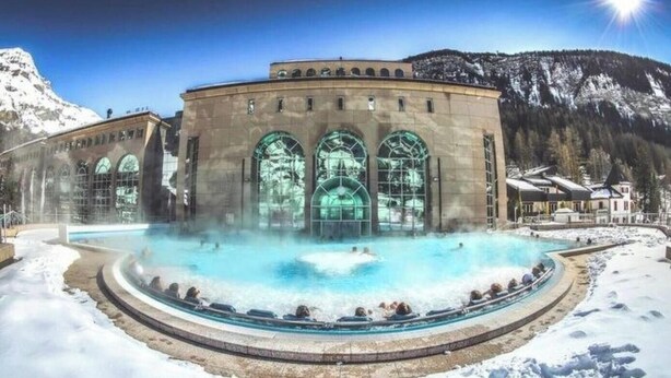 Ein Spa mit großem Außenpool, umgeben von Schnee, einem Gebäude mit Bogenfenstern und einer Bergkulisse.