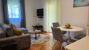 A living room with a sofa, a small round table with a vase, a dining table with chairs, and a TV mounted on the wall. - Apartment Meri 2 by Interhome (Losinj / Mali Losinj)