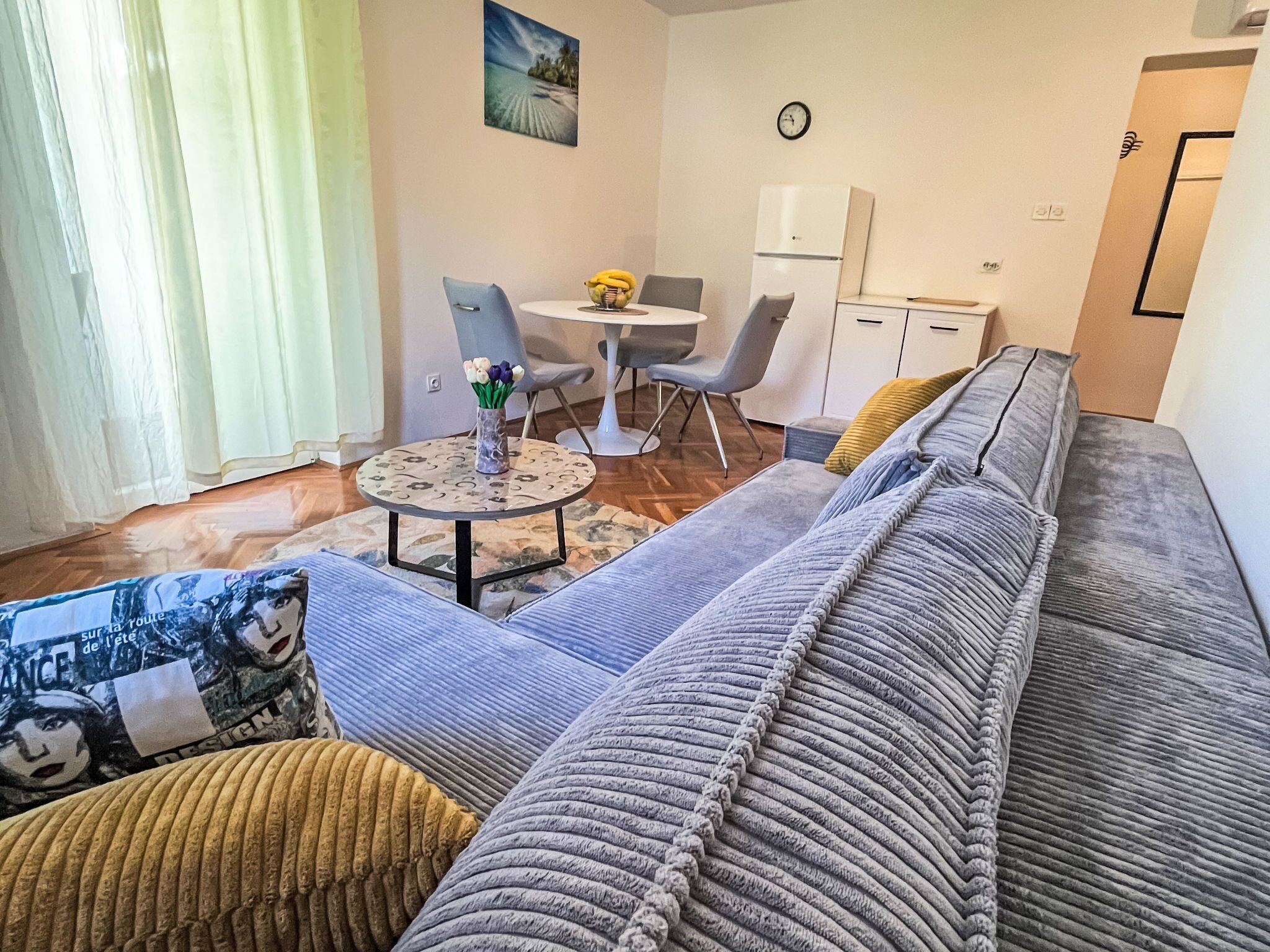 Un espace de vie comprenant un canapé, une petite table ronde avec un vase de fleurs, une table à manger avec des chaises, un réfrigérateur et une horloge murale.