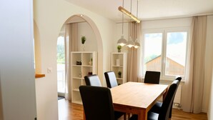 Ein Essbereich mit Holztisch und schwarzen Stühlen, einem weißen Regal mit Dekoration und einem großen Fenster mit Vorhängen.