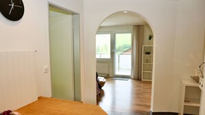 Ein Flur mit einem Holztisch, einer schwarzen Uhr, einer weißen Tür und Blick in einen Wohnbereich mit einer Couch und einem Bücherregal.