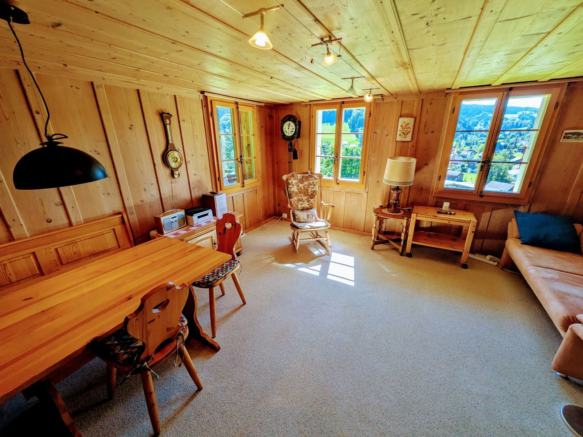Interior acogedor de una cabaña con una mesa de comedor de madera, sillas, una mecedora, un sofá, una lámpara y un reloj en la pared.