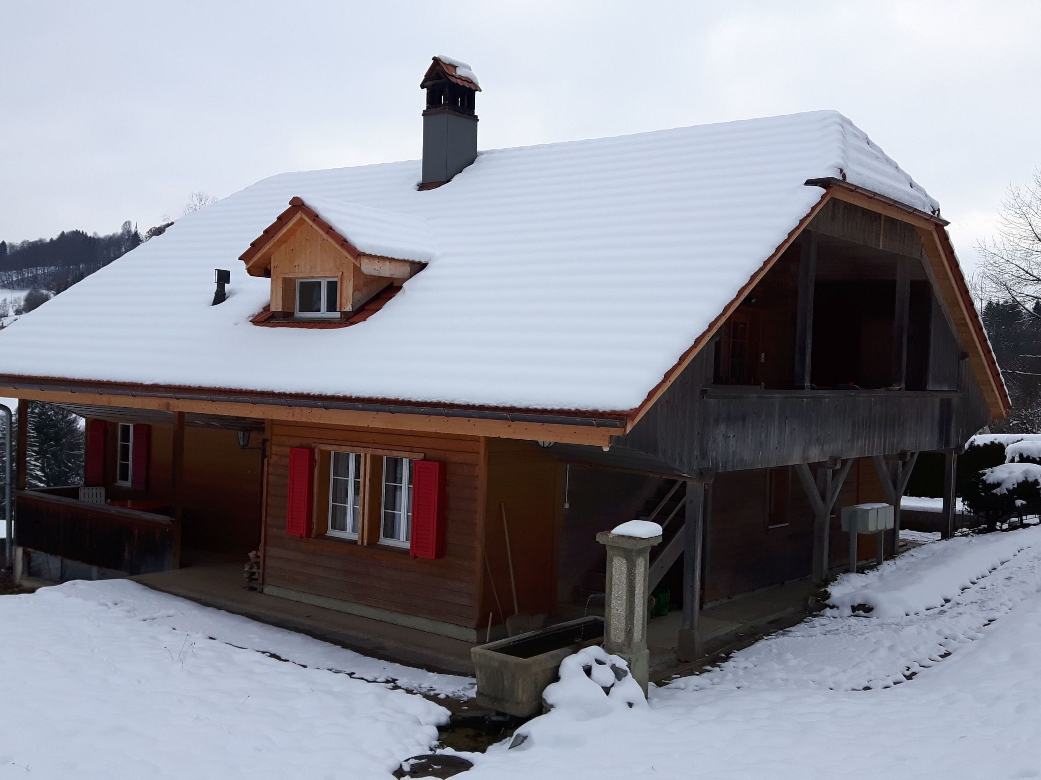 A wooden house with a steeply pitched roof covered in snow, a chimney, and a small porch area.