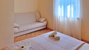 A room with a sofa, a bed with a rolled-up towel, and a window with sheer curtains.