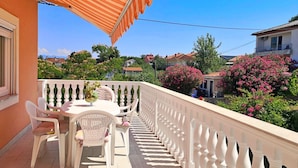 A balcony with white chairs and a table, overlooking a residential area with houses and flowering trees.