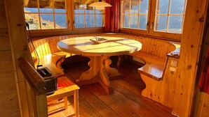 A wooden cabin interior with a round dining table, benches, and large windows offering a view of the outdoors.