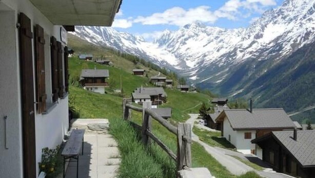 A mountain village with traditional houses, a wooden fence, and a clear view of snow-capped mountains.