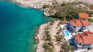Luftaufnahme einer Küstenregion mit klarem türkisblauem Wasser, felsigem Gelände und einer Ansammlung moderner Gebäude mit roten Ziegeldächern.