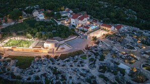 Vista aérea de un resort con cancha de tenis, varias edificaciones y jardines ajardinados.