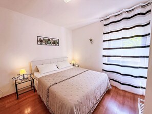 A bedroom with a bed, two bedside tables with lamps, a wall-mounted artwork, a window with striped curtains, and a small potted plant. - Apartment Zeljko by Interhome (Krk/Šilo)