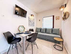 A modern living area with a sofa, a dining table with chairs, a TV mounted on the wall, and a window with blinds. - Apartment Zeljko by Interhome (Krk/Šilo)