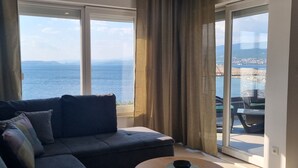 Ein Wohnzimmer mit einer Couch, einem kleinen runden Tisch mit einer Schale und großen Fenstern mit Blick auf das Meer und eine ferne Stadtlandschaft.