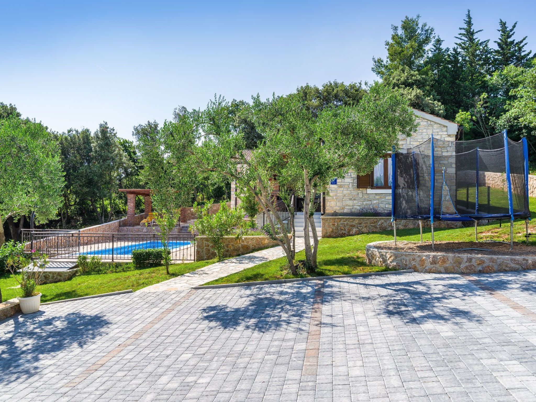 A paved outdoor area with a swimming pool, a trampoline, and a stone building.
