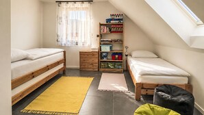 Un dormitorio pequeño con dos camas, una estantería, un ventanal con cortinas y un tragaluz.