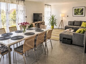 Ein modernes Wohnzimmer mit einem grauen Sofa, einem Esstisch mit Stühlen, einem Fernseher und einer Topfpflanze.