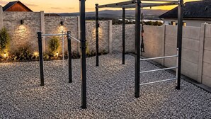 Un gimnasio al aire libre con una estructura de barra de dominadas, rodeado de suelo de grava, paredes de piedra y un atardecer de fondo.