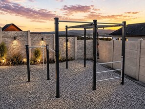 Ein Outdoor-Fitnessbereich mit einer Klimmzugstange, umgeben von Kies, Steinmauern und einem Sonnenuntergang im Hintergrund.