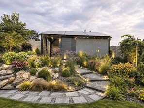 A modern building with a grey exterior, surrounded by a landscaped garden with various plants and a stone pathway. - Chalet Houbovy vrch by Interhome (Horni Plana)